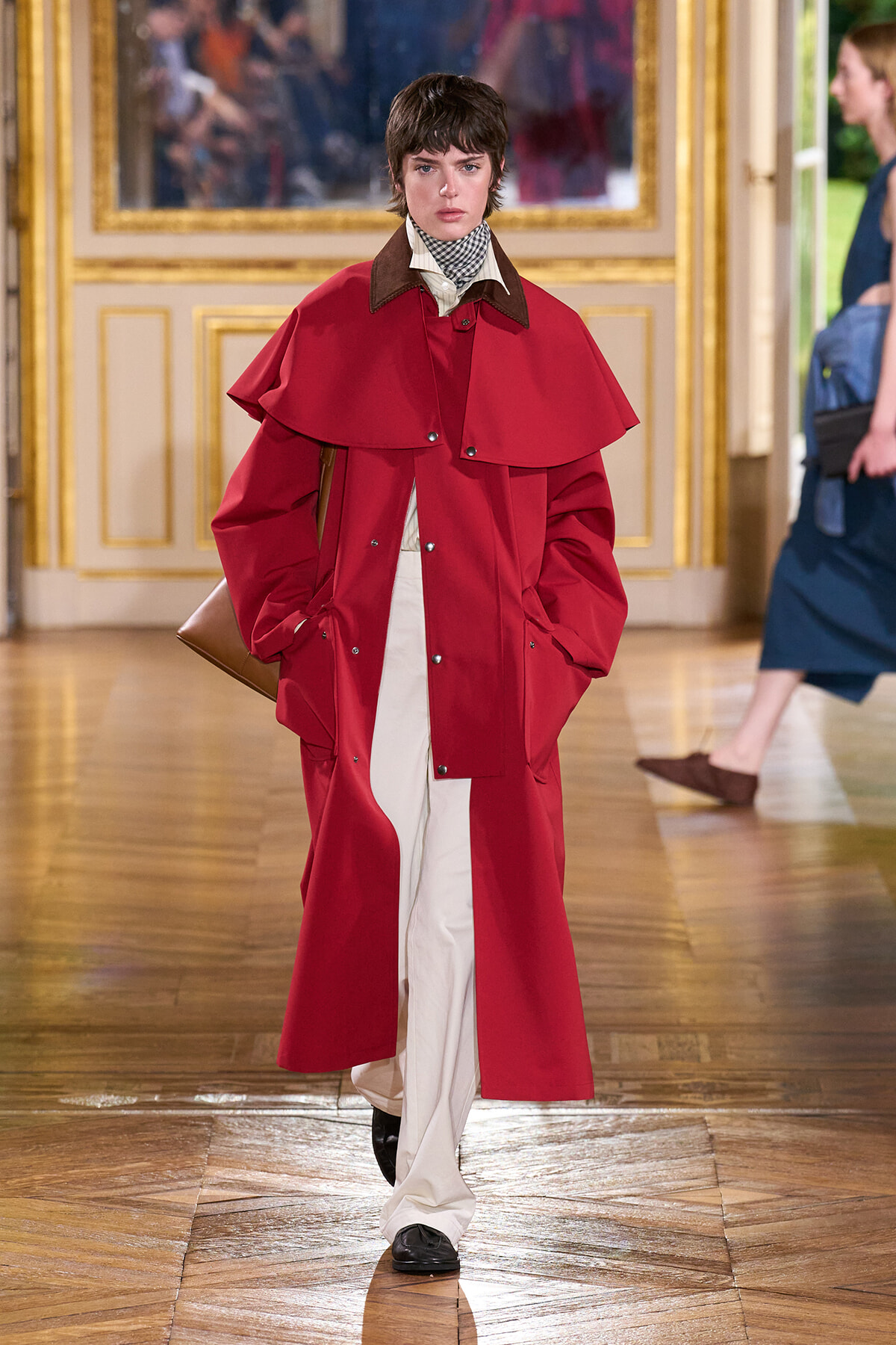 Model wearing a bright red cape-style trench over white trousers walks the runway in a gilded, ornate room.