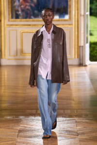 Model on a runway wearing a brown leather jacket over a pink shirt, loose blue jeans, and sandals inside a gilded room.