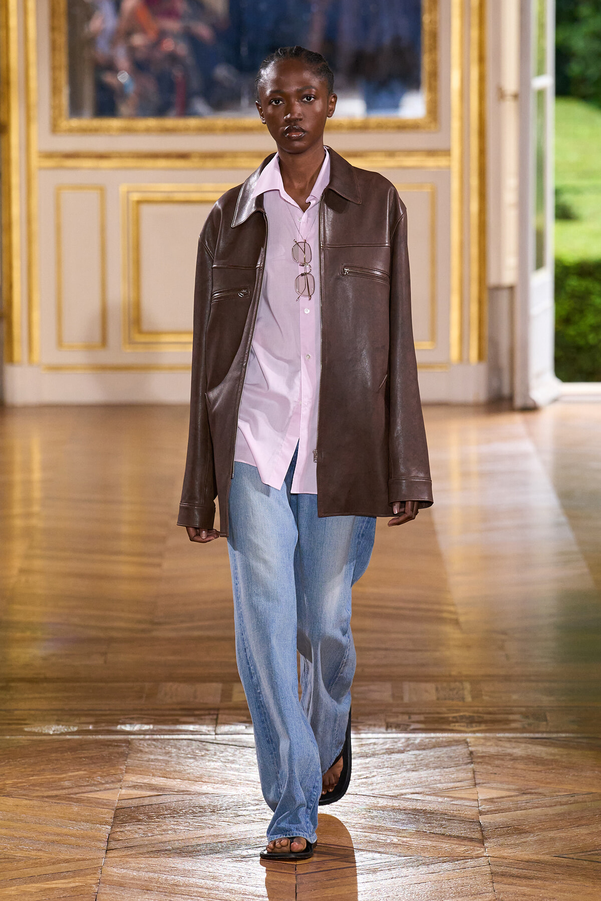 Model on a runway wearing a brown leather jacket over a pink shirt, loose blue jeans, and sandals inside a gilded room.