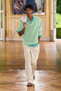 Male fashion model walking in a gilded, ornate hallway wearing a mint green sweater over a green gingham shirt and cream trousers.