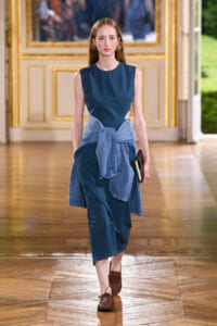 Model walks the runway in a sleeveless blue dress with a denim shirt tied at the waist, holding a small clutch.