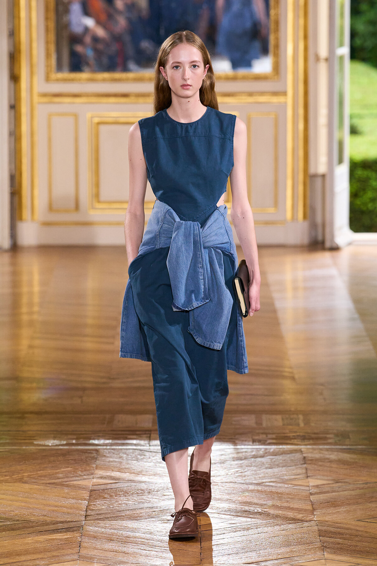 Model walks the runway in a sleeveless blue dress with a denim shirt tied at the waist, holding a small clutch.