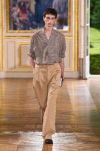 Male model walking a runway in a checked brown shirt and tan wide-leg trousers, holding a white notebook or clutch.