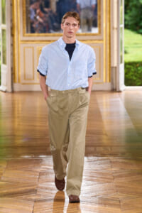 Male model walks down a gilded hallway in a light blue striped shirt over a dark tee, beige khaki pants, and brown loafers.