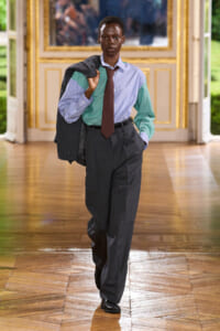 Model walking down a gilded hallway in a tailored gray suit with a blue-green striped shirt and burgundy tie, jacket slung over the shoulder.