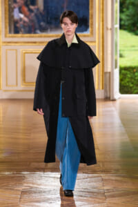 Runway model in an oversized black cape coat over a light green shirt and blue jeans in a gilded, ornate room.