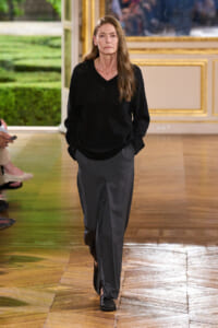 Woman with long brown hair wearing a black knit sweater and gray wide-leg pants walking down a wooden runway.