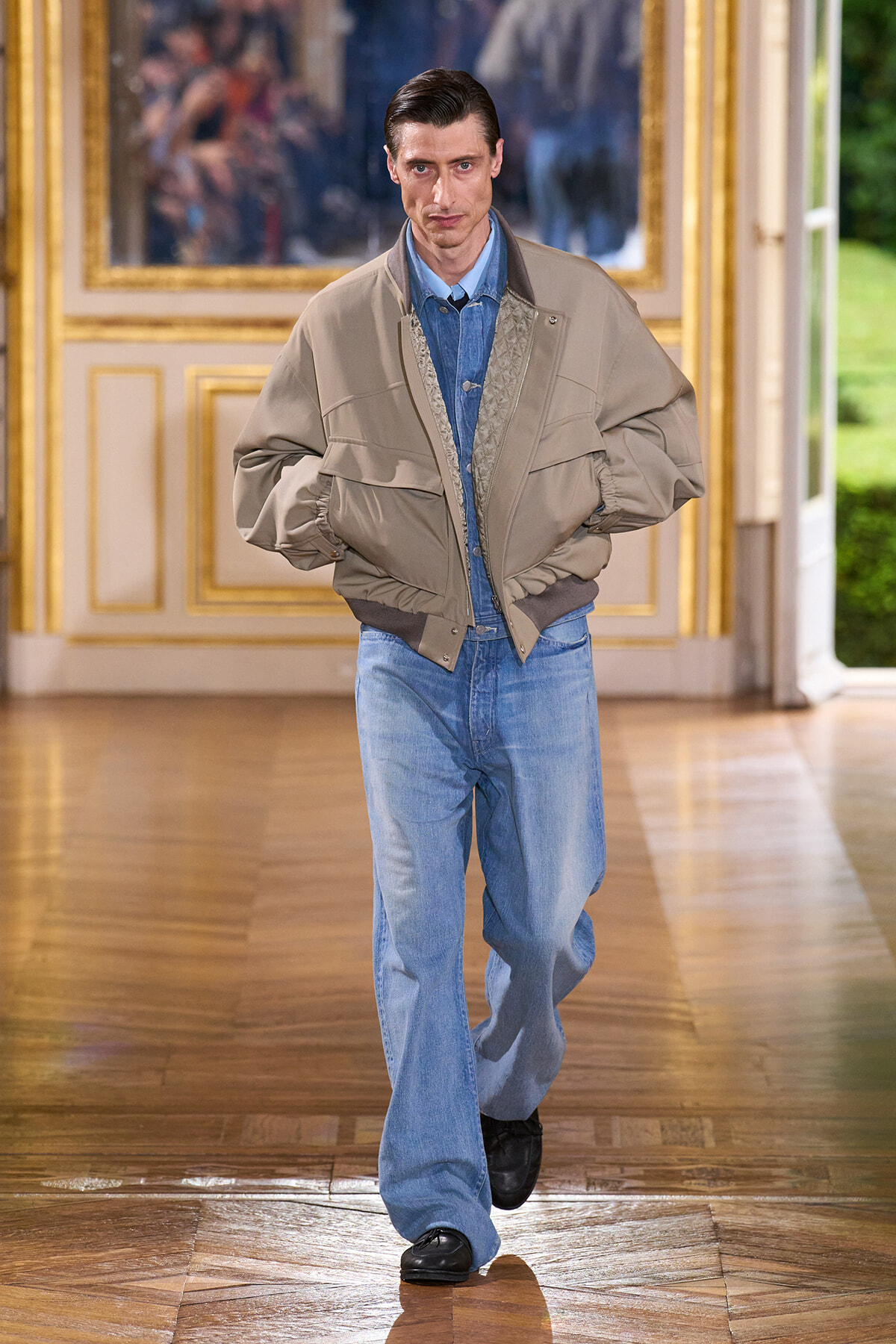 Male model walks the runway in a beige oversized bomber jacket over a blue denim shirt and light blue jeans.