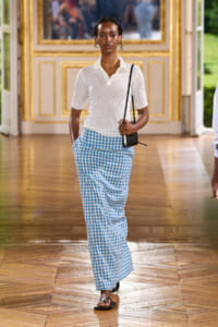 Model walking down a grand hall in a white polo and blue gingham wide-leg pants, carrying a small black crossbody bag.