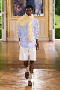 Male model walking down a gilded runway in a light blue plaid shirt, white shorts, beige scarf, and brown shoes, holding a book.