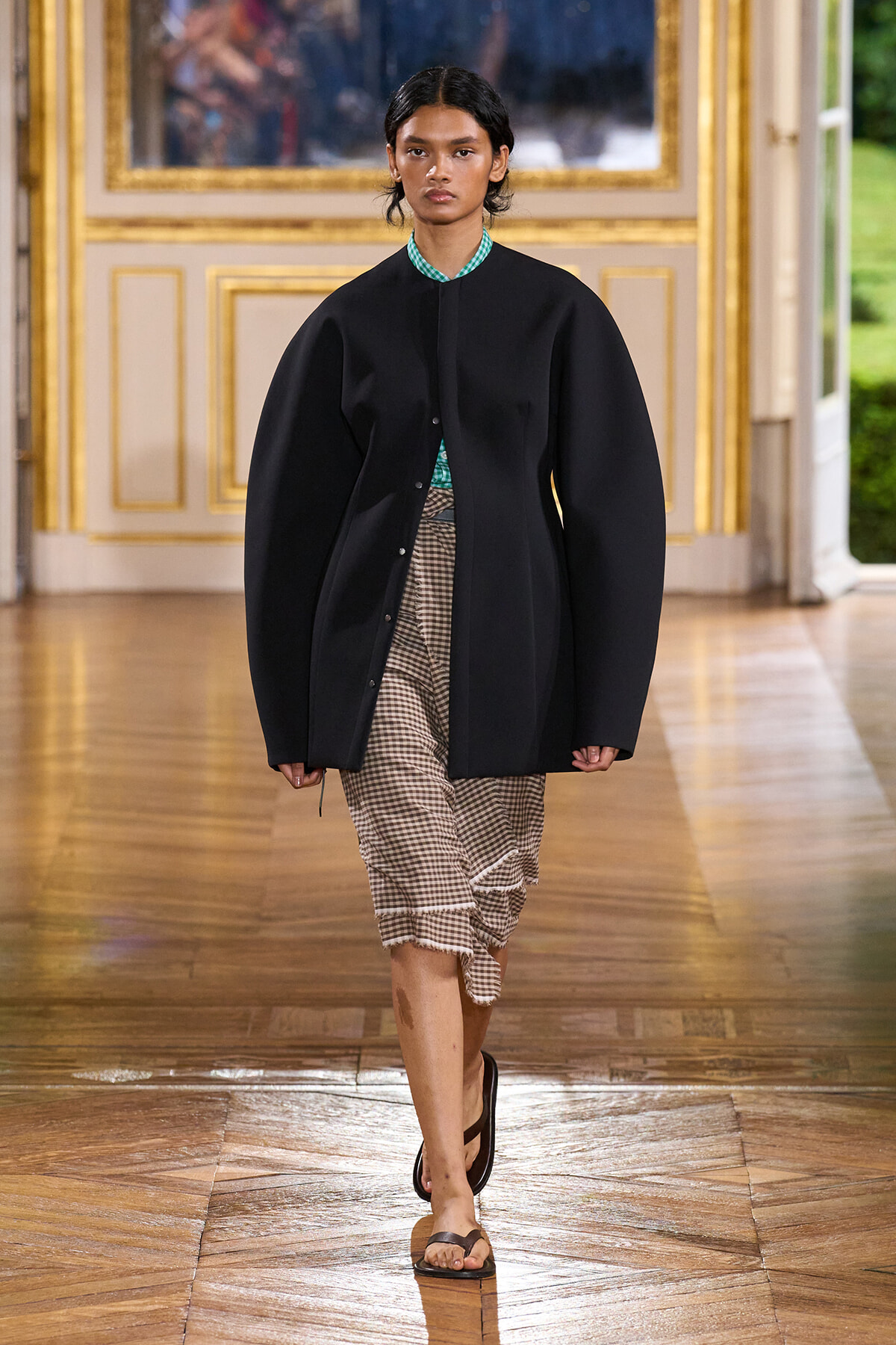 Model walks a fashion show in an oversized black coat over a green check shirt and gingham wrap skirt with fringe, in a gilded room setting.