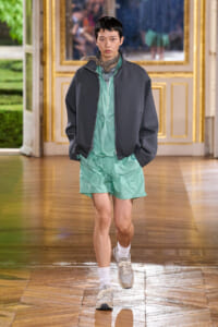 Fashion model walks a runway in mint green shorts and layered jackets in a gilded ballroom-style space.