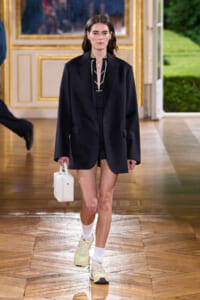 Model walking a runway in an oversized navy blazer over a black outfit, carrying a small white handbag, wearing white socks and light sneakers.