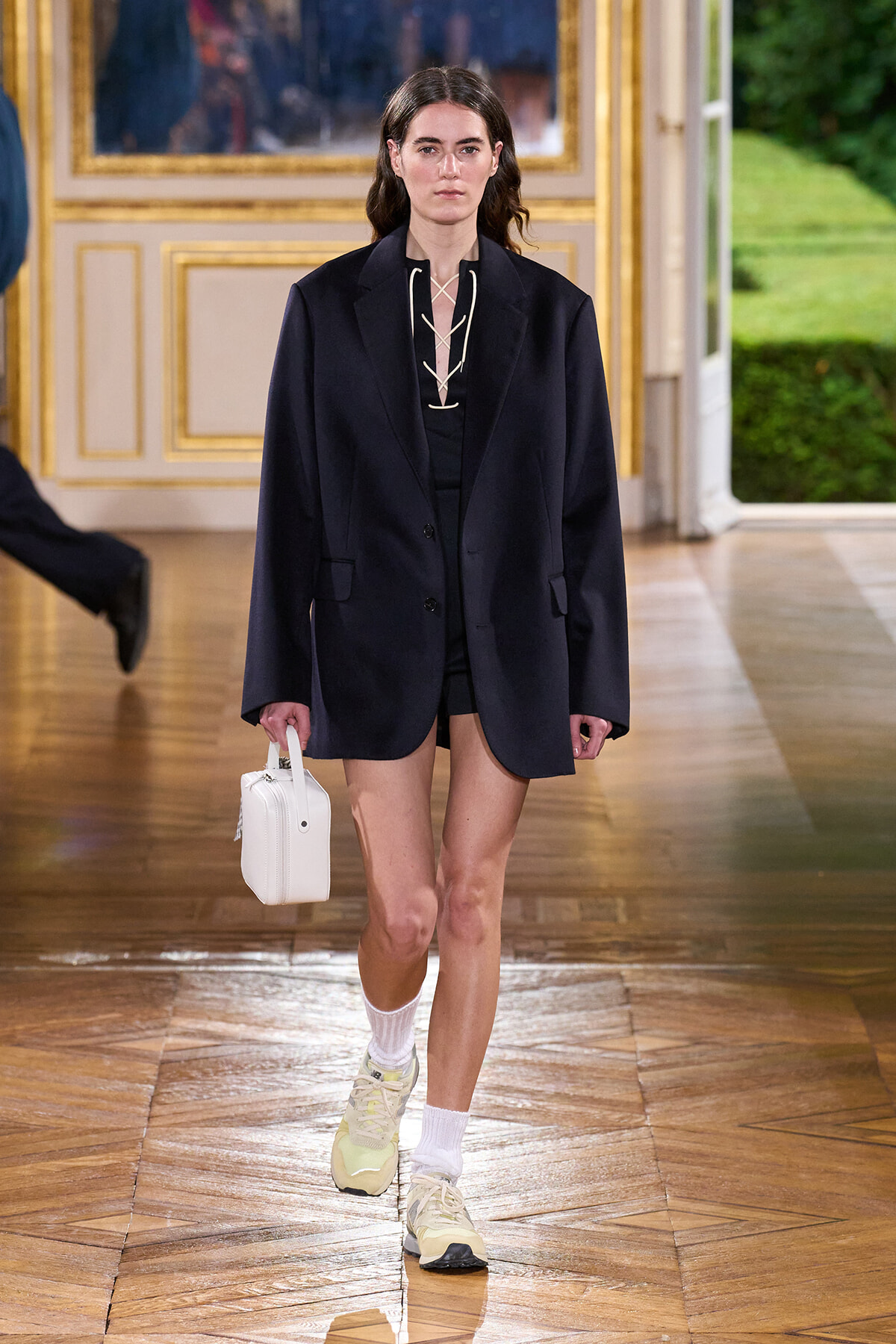 Model walking a runway in an oversized navy blazer over a black outfit, carrying a small white handbag, wearing white socks and light sneakers.