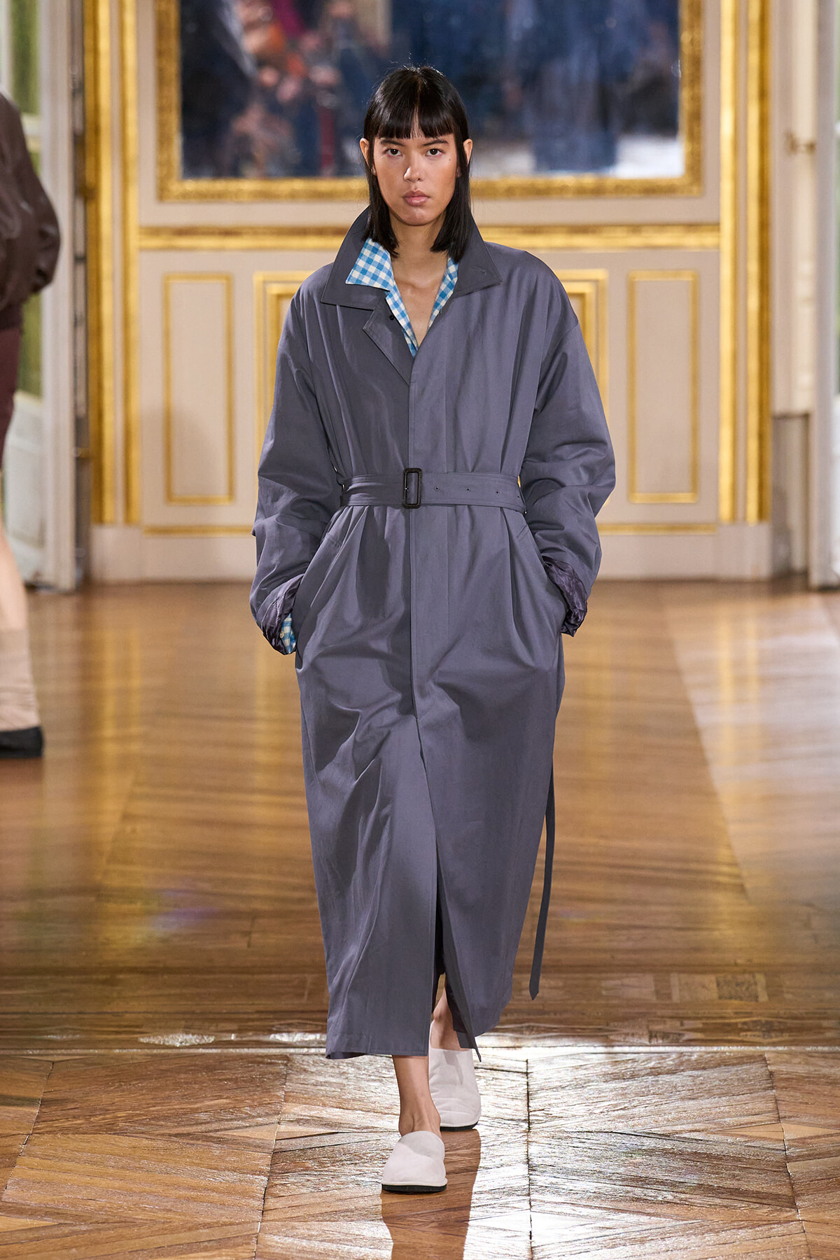 Model walking a runway in a gray belted trench coat with a blue checkered shirt collar and white slip-ons, in a gilded ballroom setting.