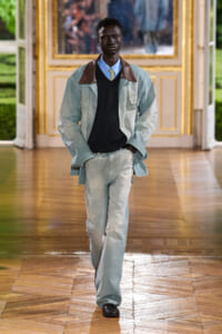 Male model walking a fashion show in a light-wash denim suit with a brown-collar jacket, blue shirt and lime tie in a gilded room setting.