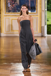 Model walking a runway in a strapless charcoal jumpsuit, carrying a gray coat and black bag in an ornate gilded room setting