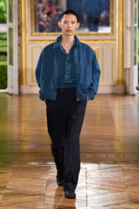 Male fashion model walks down a gilded hallway in a blue bomber jacket over a teal shirt and black trousers.