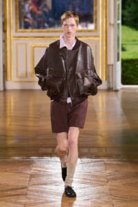 Male model on a fashion runway wearing an oversized dark brown leather bomber jacket, pale pink shirt, burgundy shorts, beige socks, and black shoes.