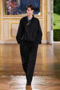 Model on a runway wearing a black oversized jacket and matching pants with a crossbody bag, in a gilded, ornate room.