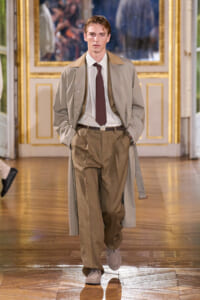 Male model walking a runway in a beige trench coat, tan trousers, white striped shirt, and burgundy tie in an ornate indoor fashion show setting