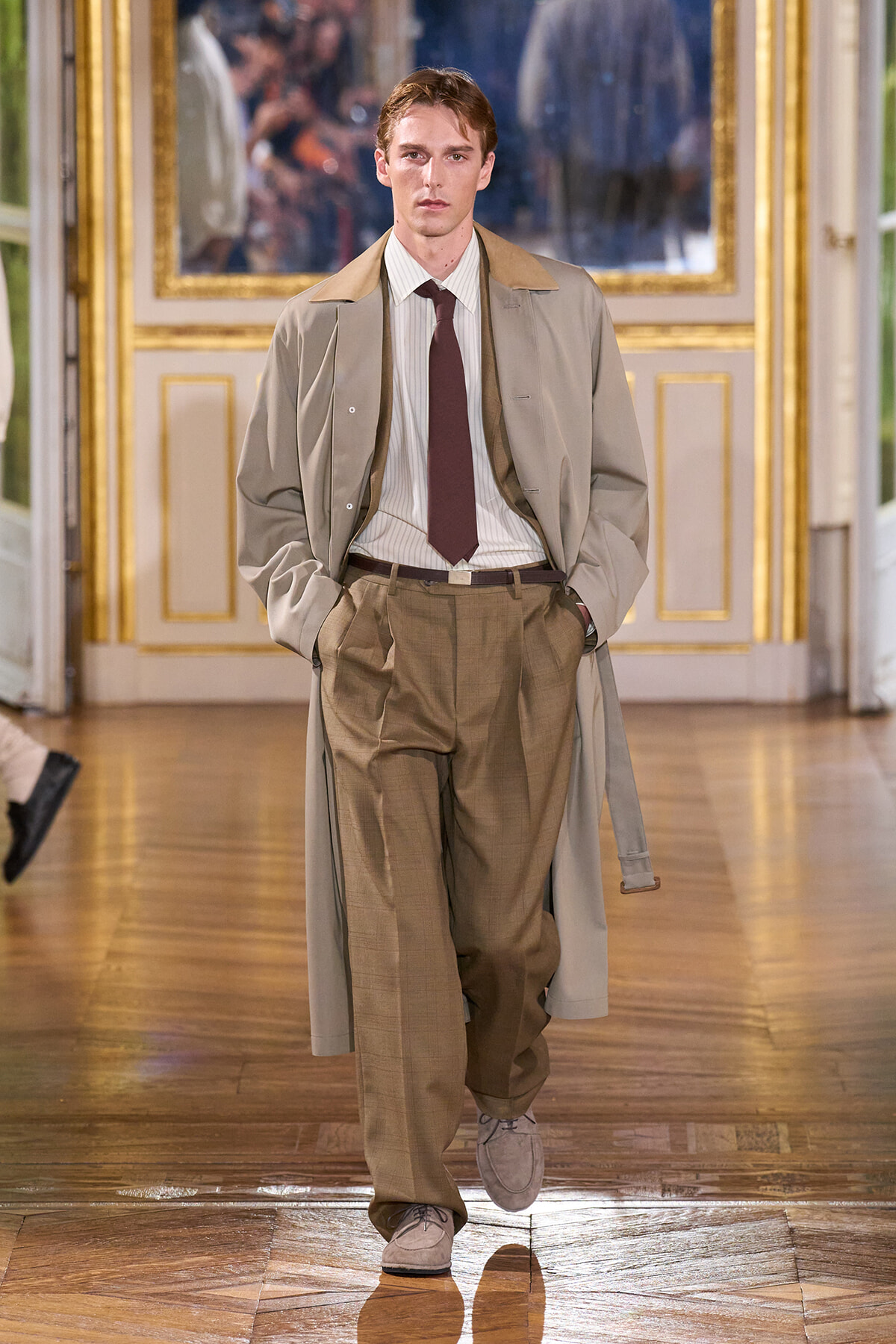 Male model walking a runway in a beige trench coat, tan trousers, white striped shirt, and burgundy tie in an ornate indoor fashion show setting