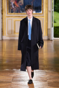 Model walking in a dark navy overcoat, white shirt, and light blue tie, carrying a small notebook in an ornate hallway.