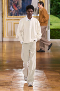Model walks a runway in a cream oversized sweater and light beige wide‑leg pants, with white wired earbuds