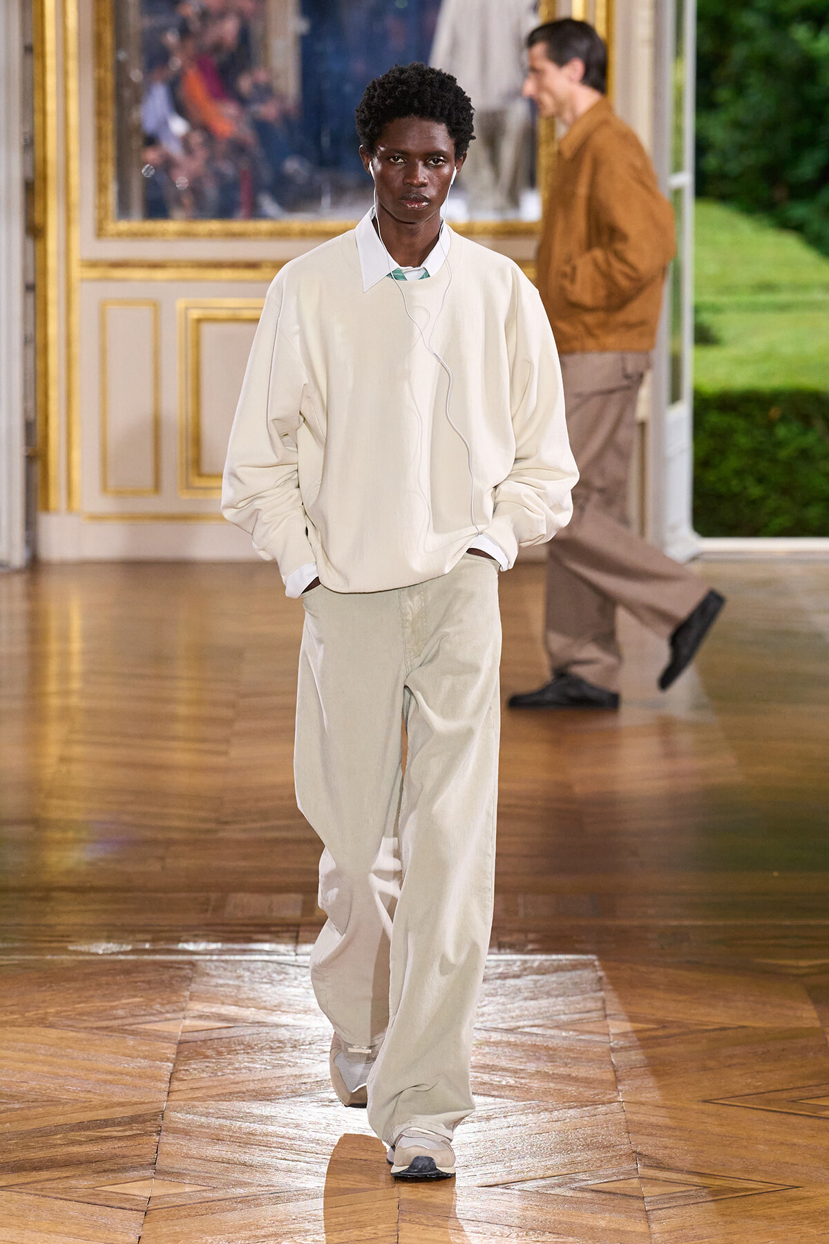 Model walks a runway in a cream oversized sweater and light beige wide‑leg pants, with white wired earbuds
