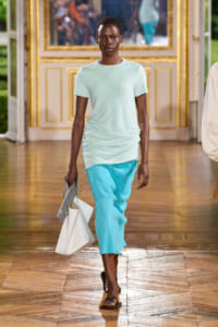 Model walks a runway in a pastel mint top and bright turquoise pants, carrying a white bag and a checkered accessory in an ornate gilded setting.