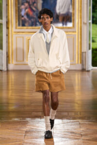 Male model walking a runway in a cream jacket, gray sweater, white shirt, tan shorts, and black shoes.