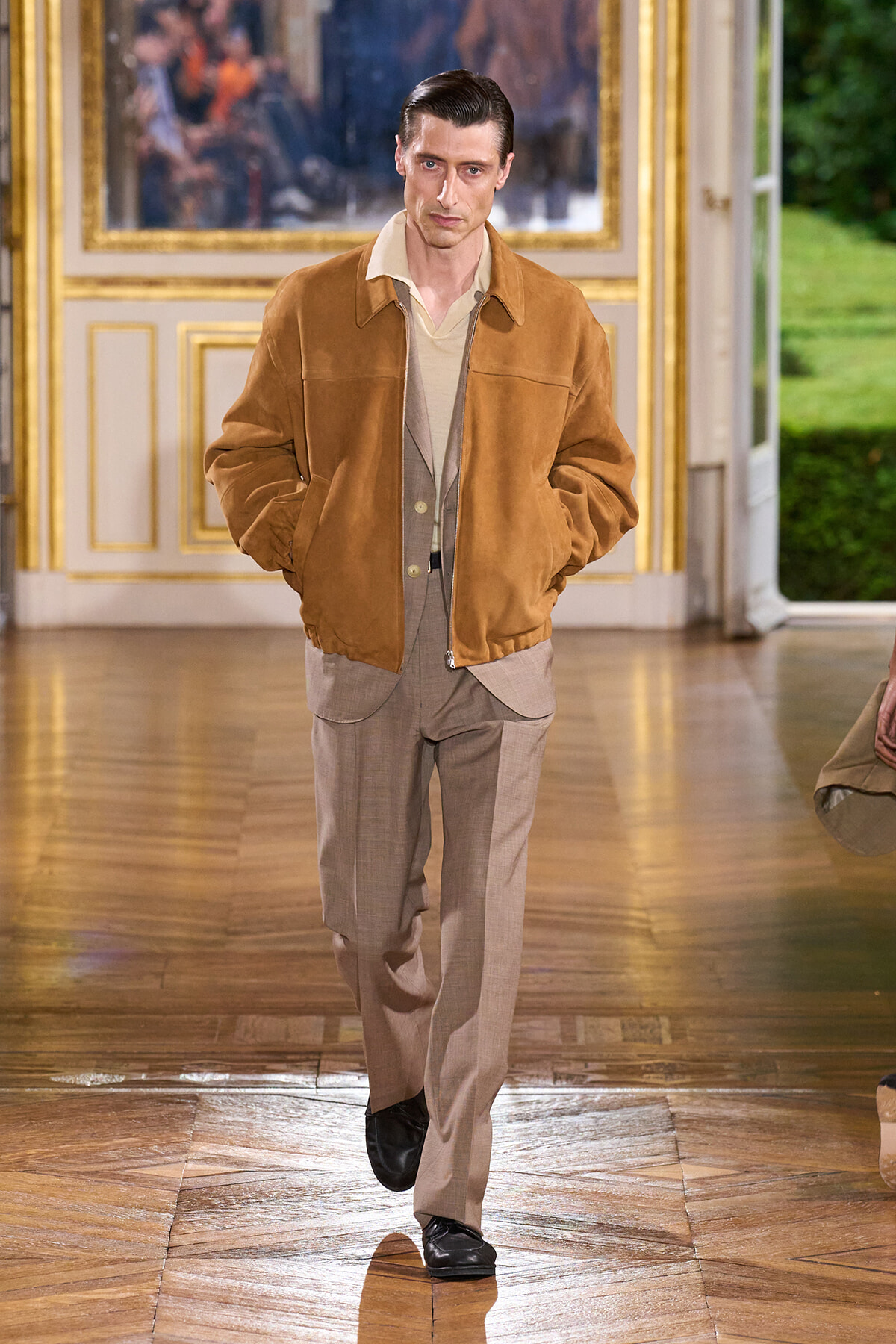Male model on a runway wearing a tan suede bomber jacket over a beige cardigan and light brown suit trousers, black shoes, in a gilded room backdrop.