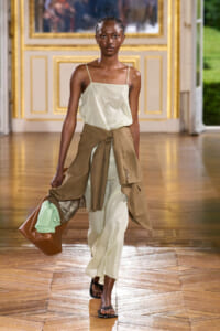 Model walks a fashion show wearing a pale green satin slip dress with thin straps, a tan blazer tied at the waist, and black sandals, carrying a large brown handbag with a mint-green sweater peeking out, on a gilded, ornate indoor runway.