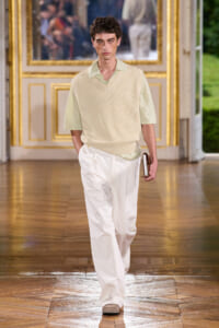 Male model walking a runway in a cream knit vest over a pale green shirt, white wide-leg pants, holding a small notebook.