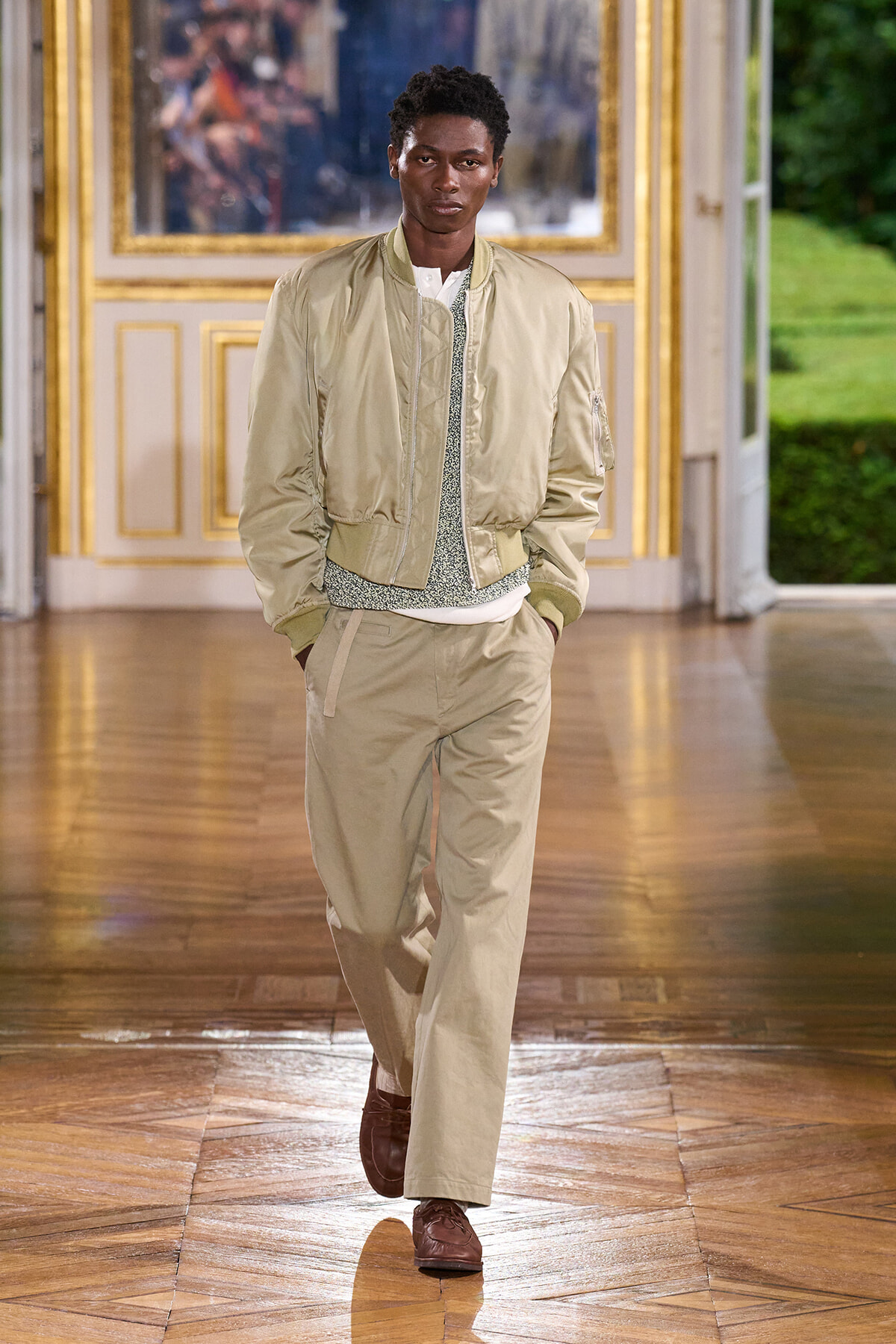 Male model walking a runway in a beige bomber jacket and matching pants inside a gilded, ornate room; brown loafers.