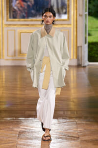 Model on a runway in a mint-green oversized coat with tan collar, cream knit top, white wide-leg pants, and a yellow scarf-belt.