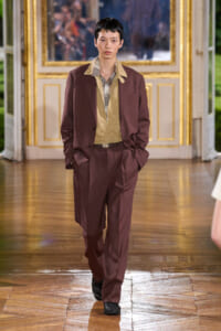 Modelling a burgundy suit with a tan inner shirt, walking down a wooden runway in a grand, gilded room.