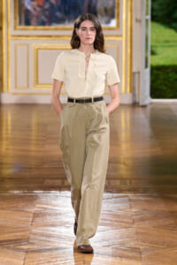 Model walking a runway in a cream blouse with lace-up front and high-waisted khaki trousers with a black belt and brown shoes in an ornate gold-trimmed room.