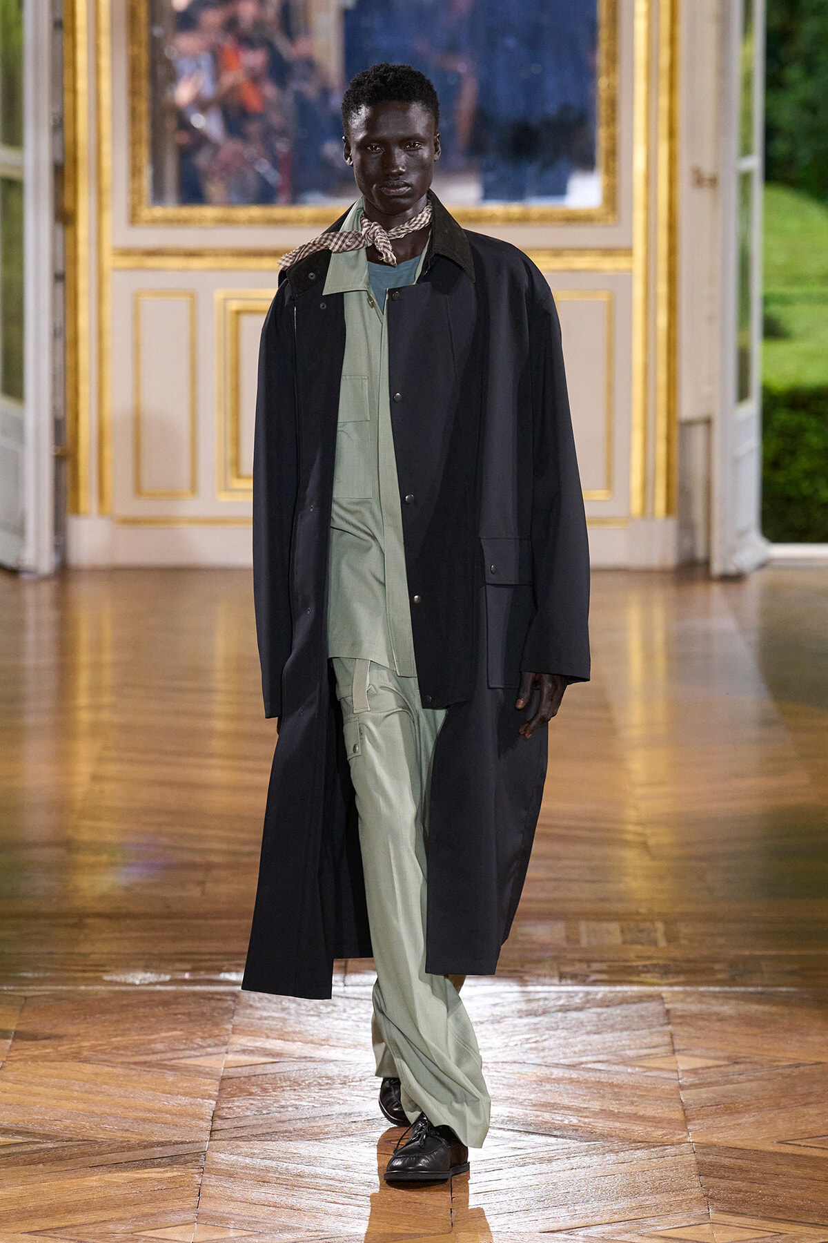 Male model walking a fashion runway in a long black coat over a sage-green utility jumpsuit, with a patterned scarf at the neck.