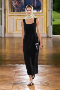 Model walking a runway in a black sleeveless midi dress, beige headscarf, and black clutch in a gilded interior.