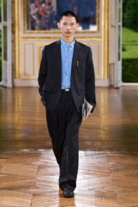 Male model walking down a runway in a dark pinstripe suit and light blue shirt, holding papers in his left hand.