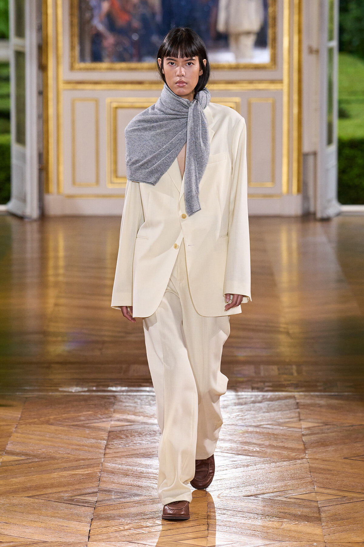 Model walking a runway in a loose cream suit with an oversized blazer and matching wide-leg pants, accessorized with a gray scarf wrapped around the neck in a draped knot, brown shoes, and a gilded, ornate backdrop behind a polished wooden floor.