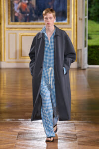 Male model walking a fashion runway wearing blue-and-white checkered loungewear and a long dark overcoat in a gilded room setting.