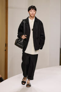 Model on a runway wearing a black oversized jacket over a cream button-down shirt, black cropped pants, strappy black sandals, and a black shoulder bag.