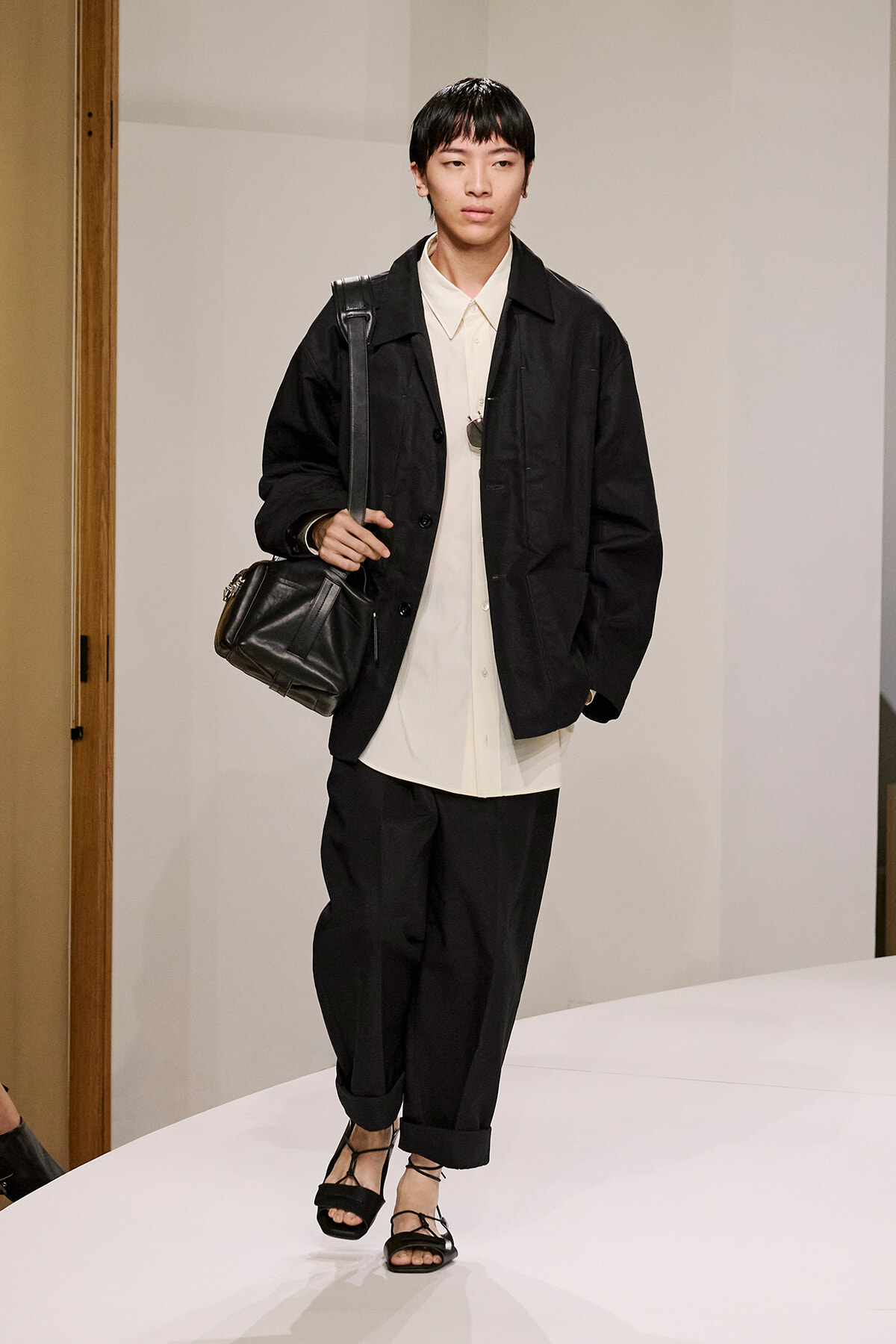 Model on a runway wearing a black oversized jacket over a cream button-down shirt, black cropped pants, strappy black sandals, and a black shoulder bag.