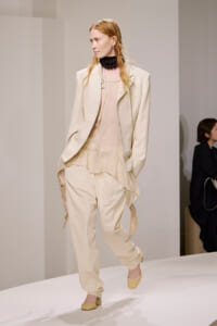 Model on a runway wearing a cream-colored layered suit with a blazer, draped top, matching pants, and a chunky black beaded necklace.