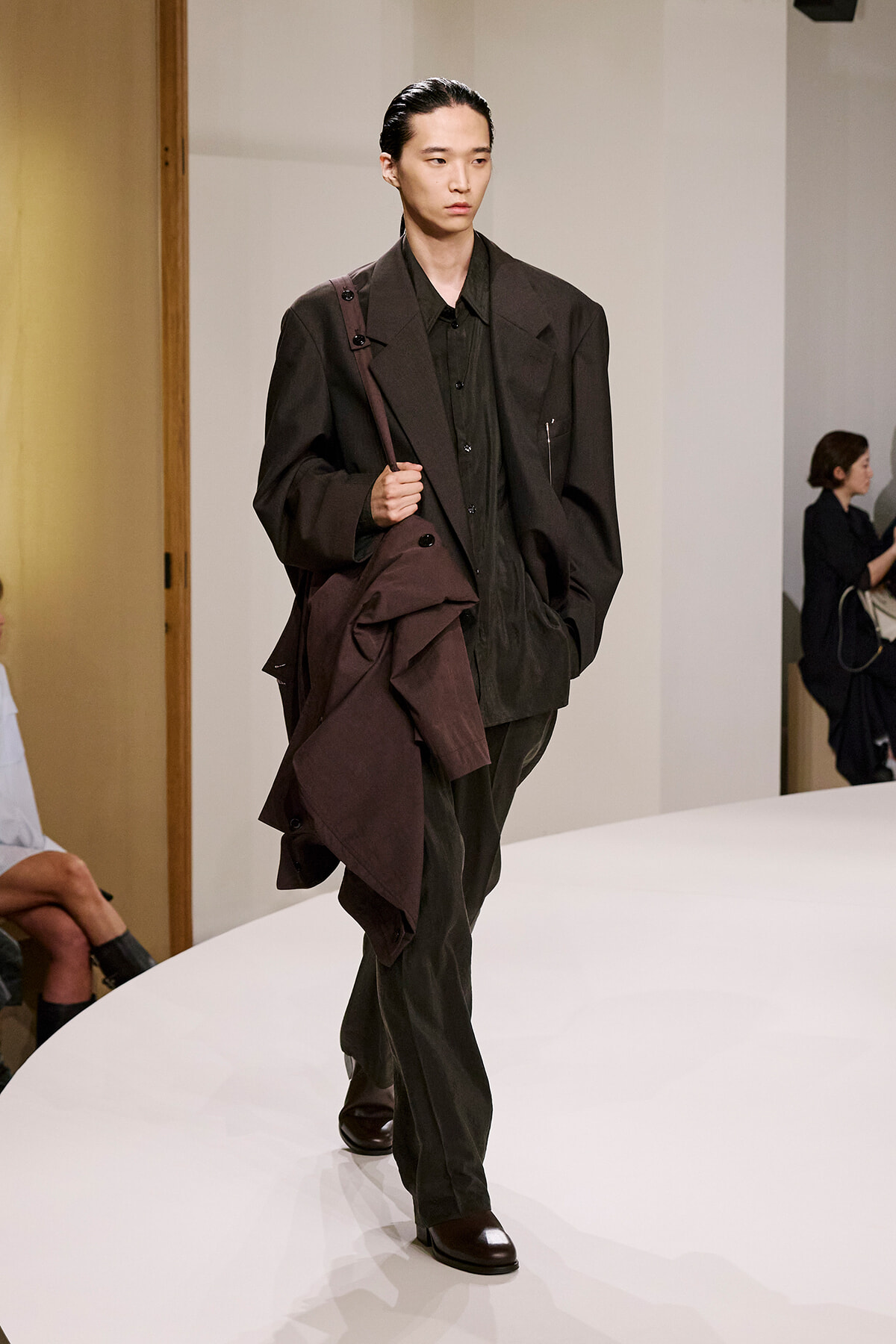 Male model walks the runway in an oversized dark suit, brown coat slung over his shoulder with a bag strap across his chest.