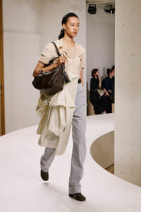 Model walking a white curved runway wearing a beige layered trench coat over light gray jeans, carrying a large brown handbag on her shoulder.