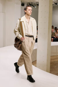 Male model walks a white runway in a cream/beige suit with a matching shirt, black belt, black boots, and a large brown shoulder bag.