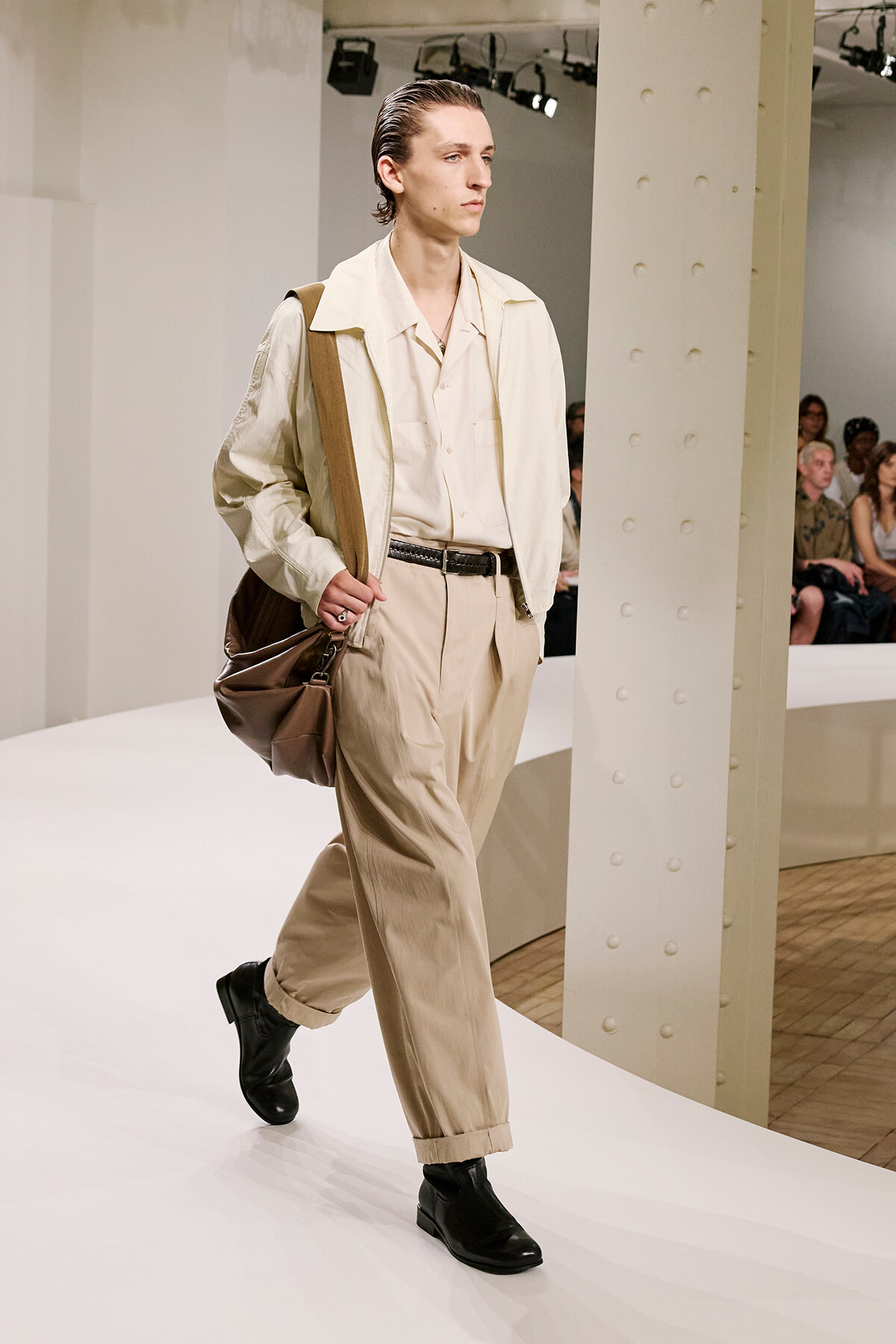 Male model walks a white runway in a cream/beige suit with a matching shirt, black belt, black boots, and a large brown shoulder bag.